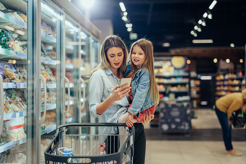Mother shopping with daughter in grocery store, checking smartphone for Complex Community Federal Credit Union mobile banking updates.